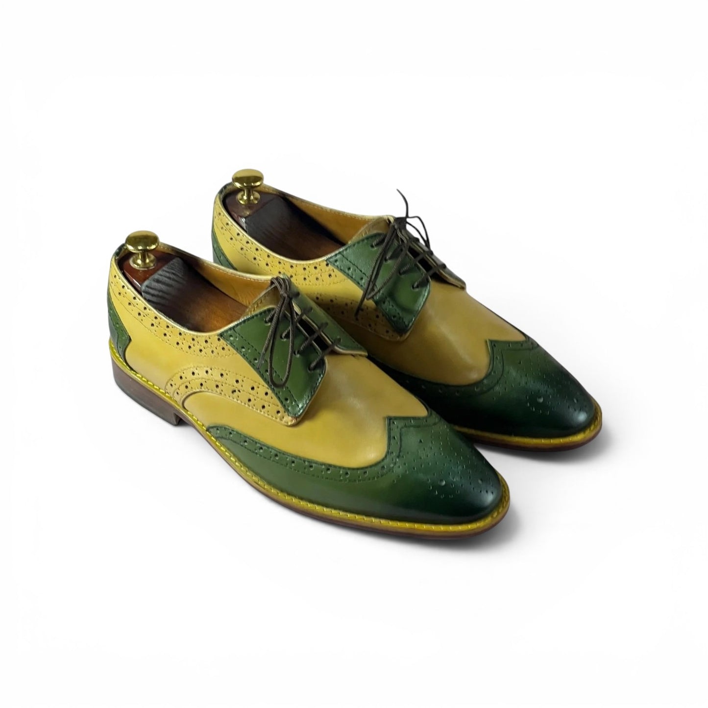 A pair of men's wingtip oxford shoes in a two-tone yellow and dark green color-blocked design, displayed on wooden shoe trees against a plain white background.