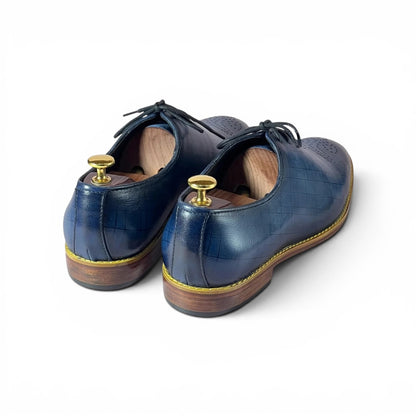 A pair of navy blue crocodile-embossed leather oxford shoes with black laces and gold-toned metal shoe trees inside, shown from a rear three-quarter angle on a white background.