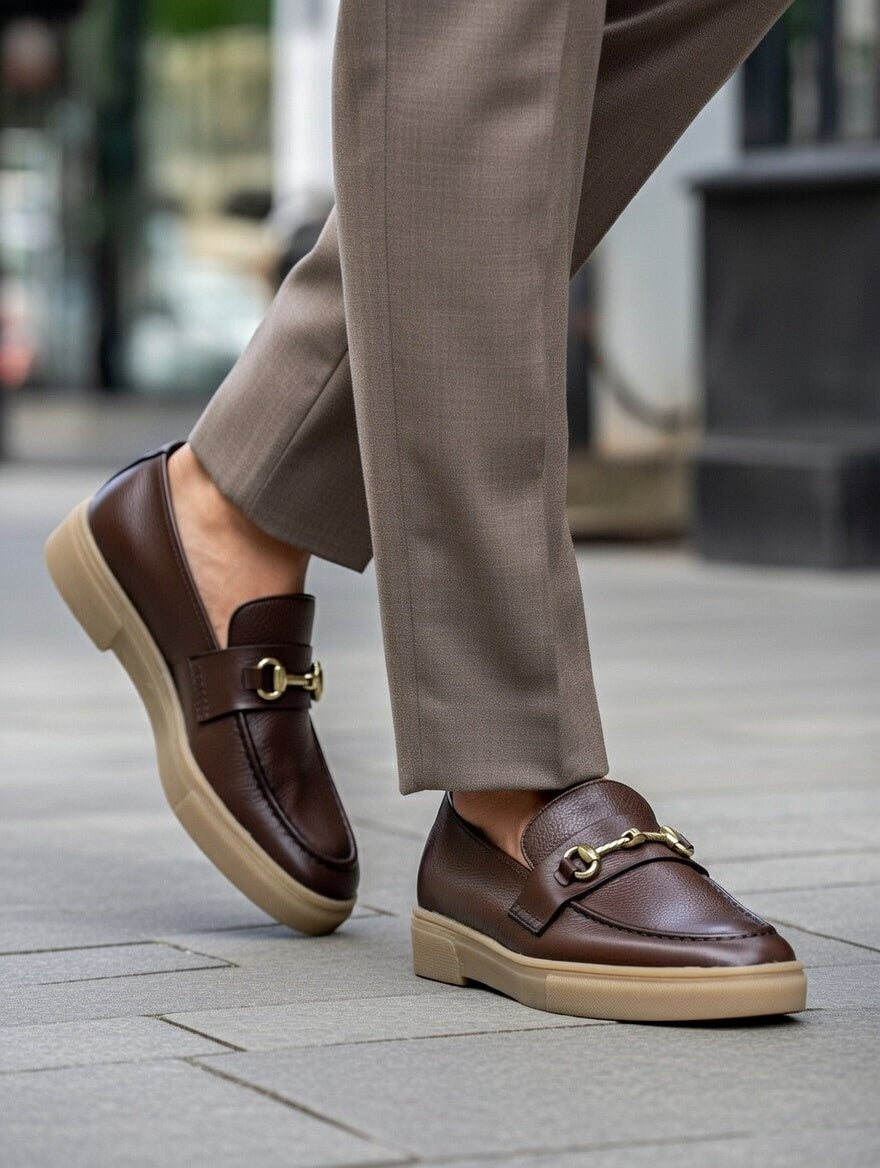 Brown loafers with gold buckles worn with beige pants on a blurred street background