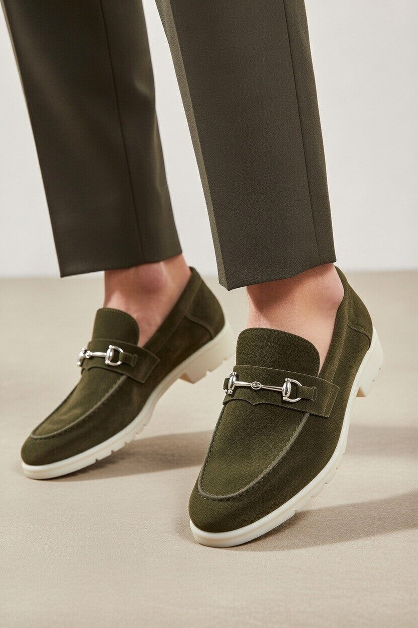 Green loafers with silver buckles worn with dark green pants on a neutral background