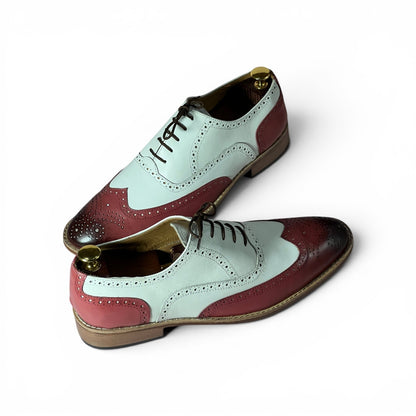 A pair of men's wingtip oxford shoes with a color-blocked design featuring pale mint green, dusty rose pink, and dark burgundy panels, finished with black laces and gold-tone metal shoe trees inside.