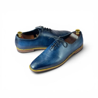 A pair of men's blue leather oxford shoes with brogue detailing, displayed on wooden shoe trees against a clean white background.