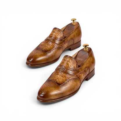 A pair of men's brown leather brogue loafers with embossed floral detailing on the toe, displayed on wooden shoe trees against a clean white background.