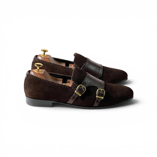 A pair of dark brown suede and leather double strap loafers with gold buckle details, displayed on wooden shoe trees against a white background.
