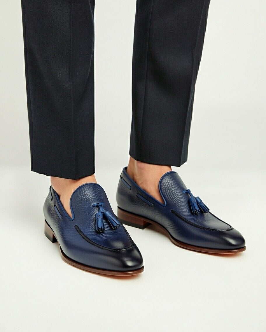 Blue loafers with tassels worn with black pants on a light gray background