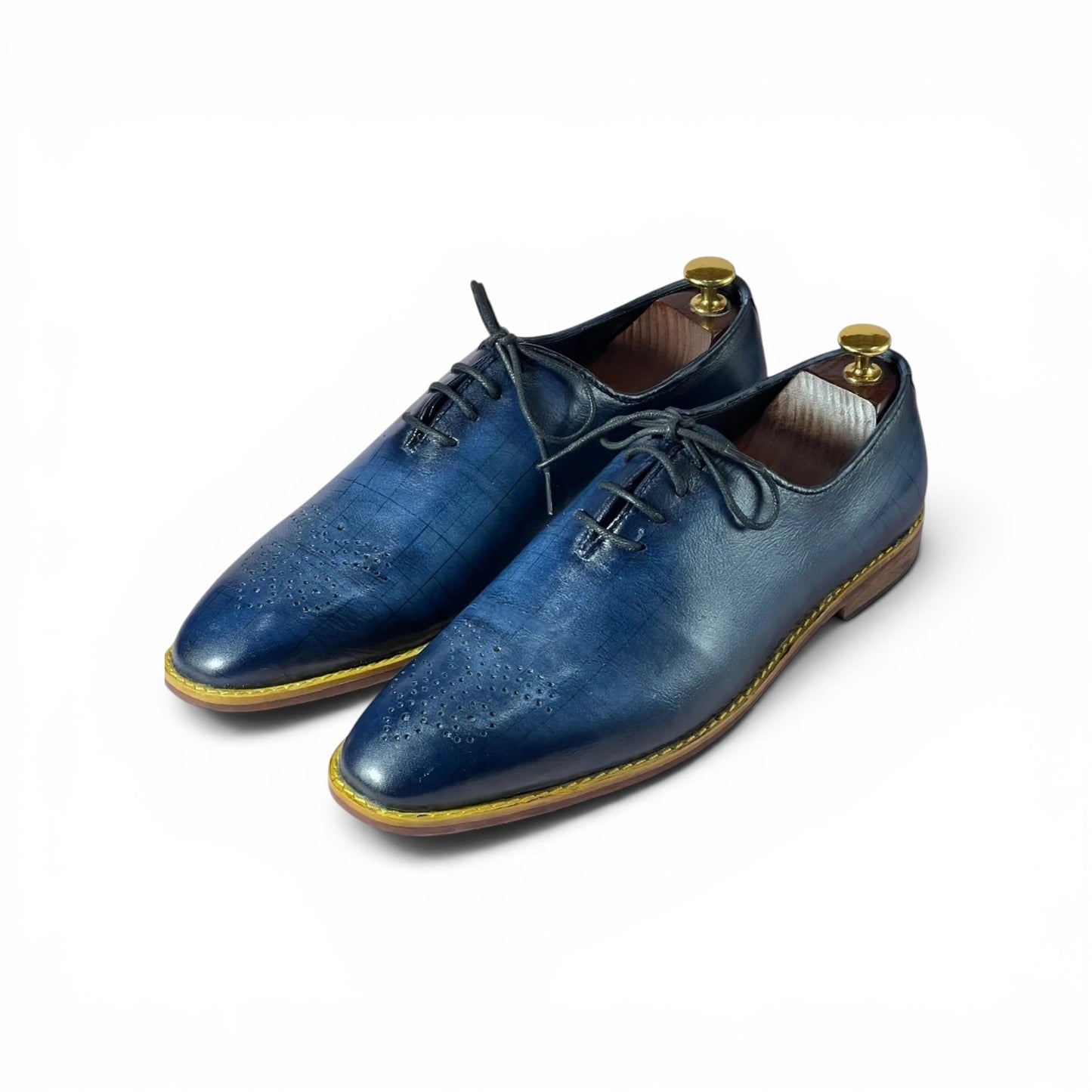 A pair of men's blue leather oxford shoes with a subtle embossed pattern, black laces, and a light brown sole with a gold welt stitch, displayed on a white background with shoe trees inside.