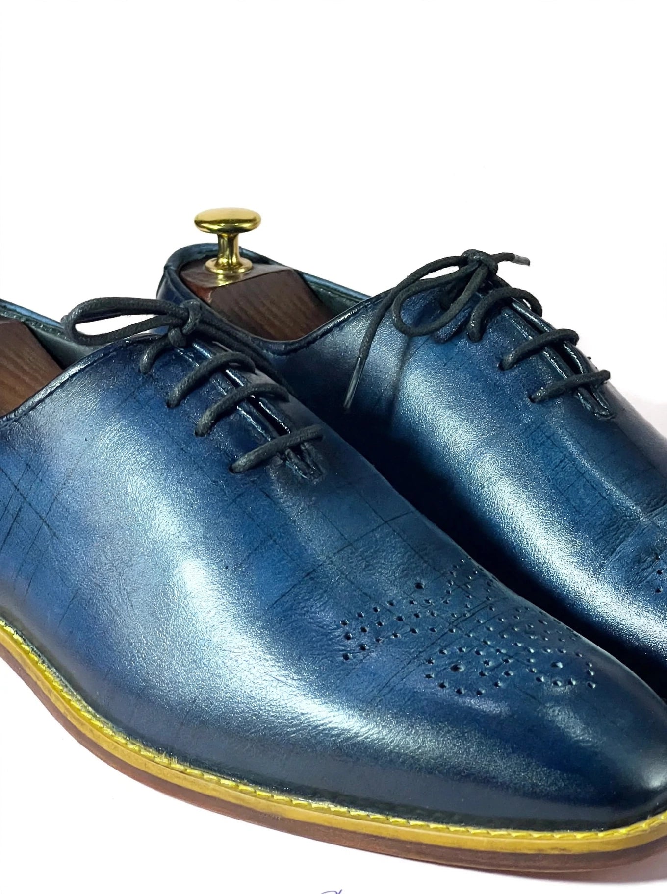 A pair of men's blue leather oxford shoes with black laces and a yellow welt stitch, displayed on wooden shoe trees against a white background.
