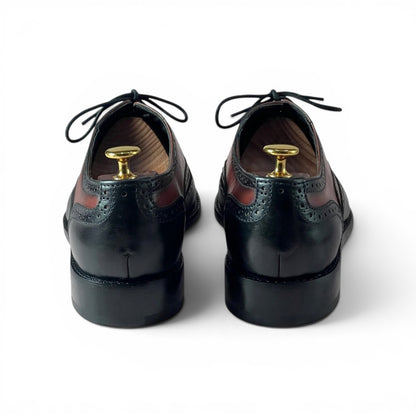 A pair of black leather wingtip oxford shoes shown from the back, with gold shoe tree inserts in the heels, on a clean white background.
