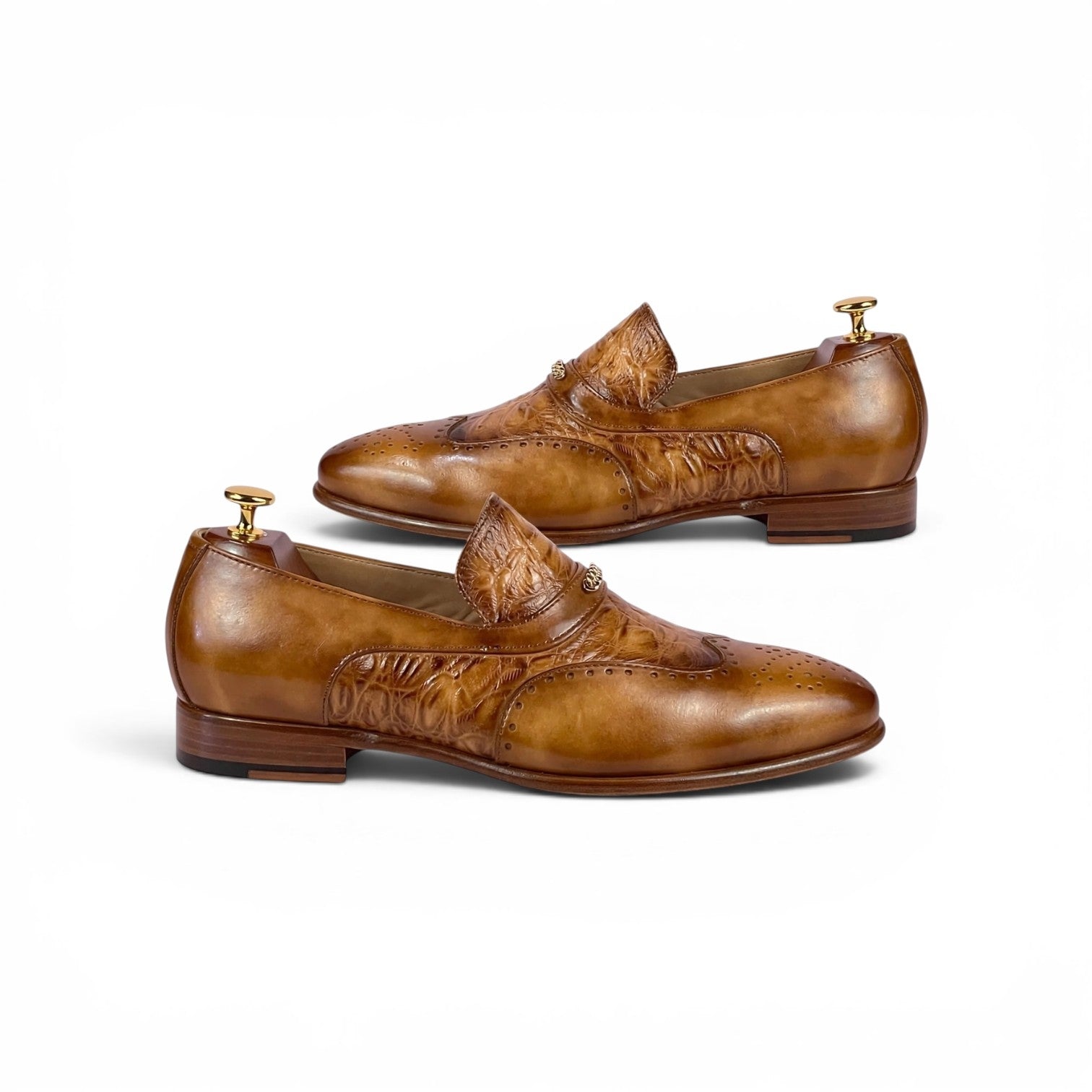 A pair of brown leather penny loafers with embossed floral detailing, shown on wooden shoe trees against a plain white background.