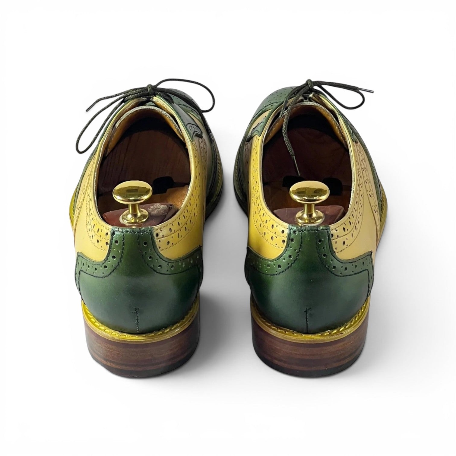 A pair of men's wingtip oxford shoes shown from the back, featuring a color-blocked design with dark green, mustard yellow, and brown leather panels, displayed on wooden shoe trees against a white background.