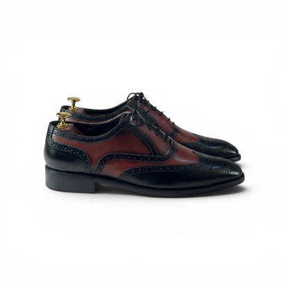 A pair of men's wingtip oxford shoes in black and dark red leather, displayed on wooden shoe trees with gold knobs against a plain white background.