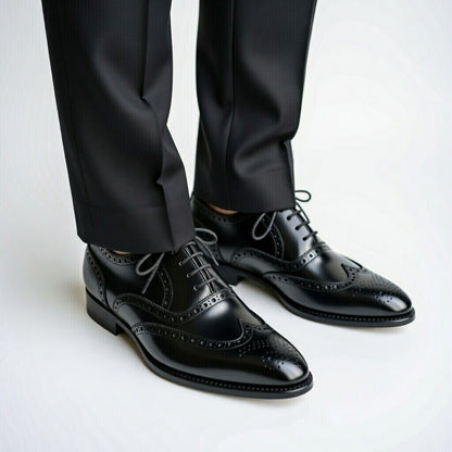 Black dress shoes with wingtip design worn with black pants on a light gray background