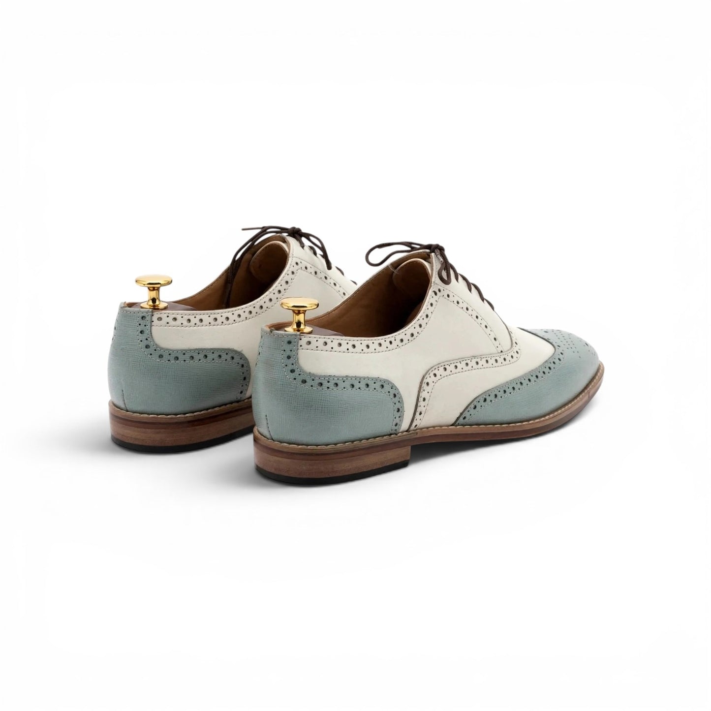 A pair of men's wingtip oxford shoes in a two-tone colorway of light blue and off-white, shown from a rear three-quarter angle on a clean white background. The shoes are displayed on wooden shoe trees with gold-colored caps.