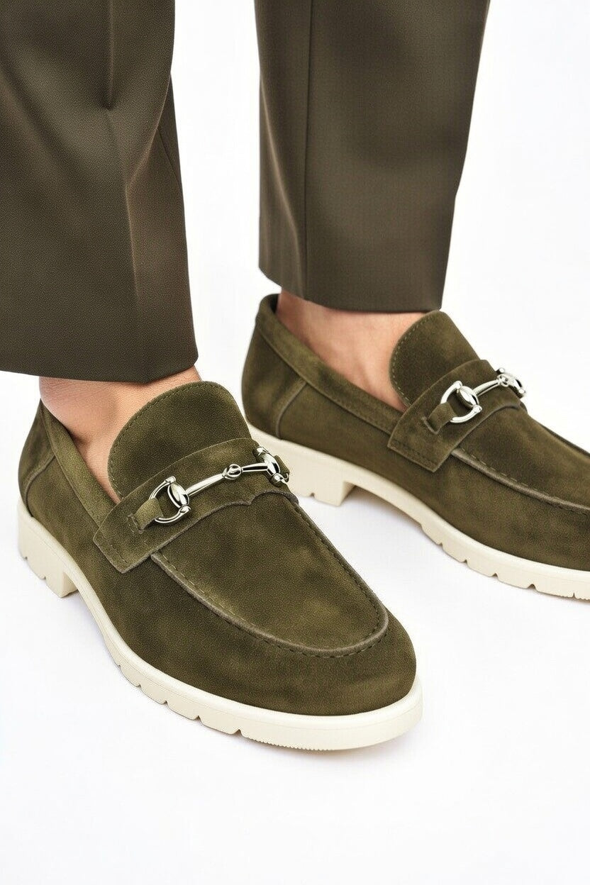 Green suede loafers with a silver buckle worn with olive green pants on a white background