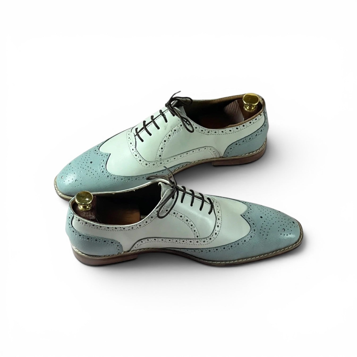 A pair of men's wingtip oxford shoes in a two-tone light blue and white colorway, featuring black laces and brass shoe trees inside, set against a plain white background.