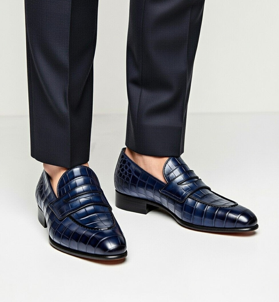 Navy blue crocodile-textured loafers worn with black pants on a light gray background