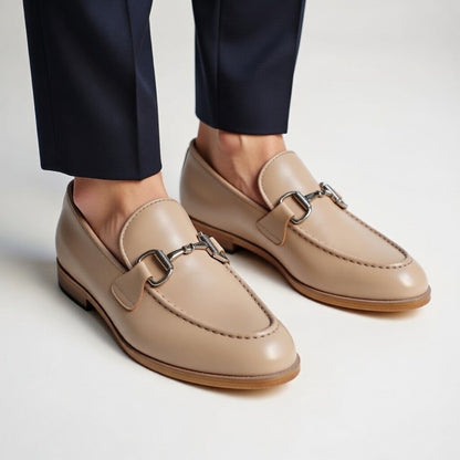 Beige loafers with metallic buckles worn with navy pants on a light background