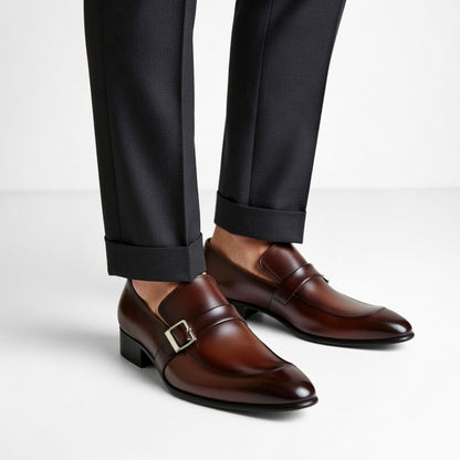 Brown dress shoes with a silver buckle worn with black pants on a light gray background