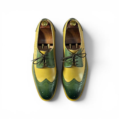 A pair of men's wingtip oxford shoes in a distinctive yellow and green color-blocked design, displayed on wooden shoe trees against a clean white background.
