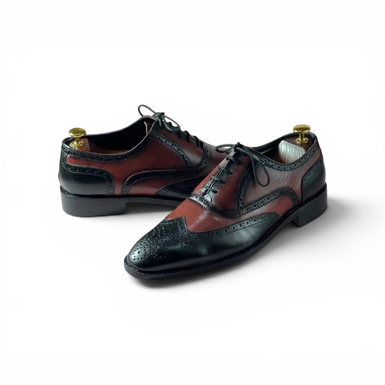 A pair of men's wingtip oxford shoes with a dark brown and black brogue design, displayed on brass shoe trees against a white background.