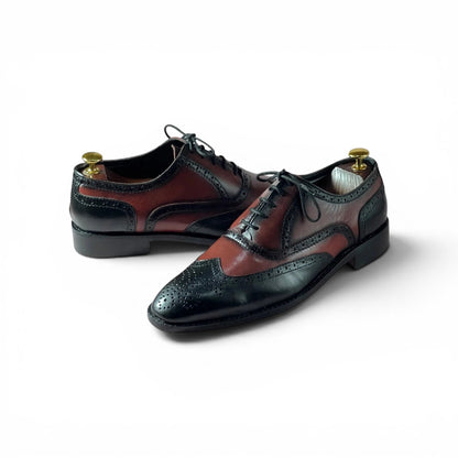 A pair of men's wingtip oxford shoes with a dark brown and black brogue design, displayed on brass shoe trees against a white background.