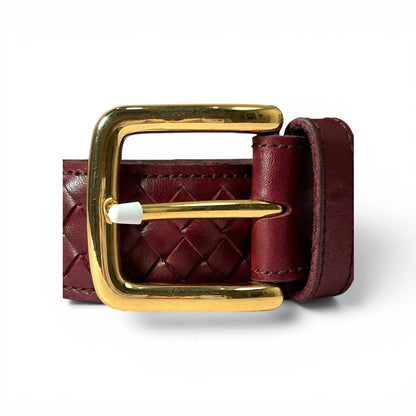 A close-up of a woven leather belt in burgundy with a polished gold buckle and a white tip on the metal pin, set against a plain white background.