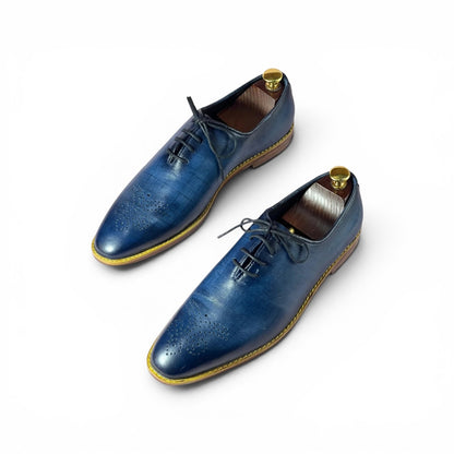 A pair of men's blue leather oxford shoes with brogue detailing, shown on white background with wooden shoe trees inside.