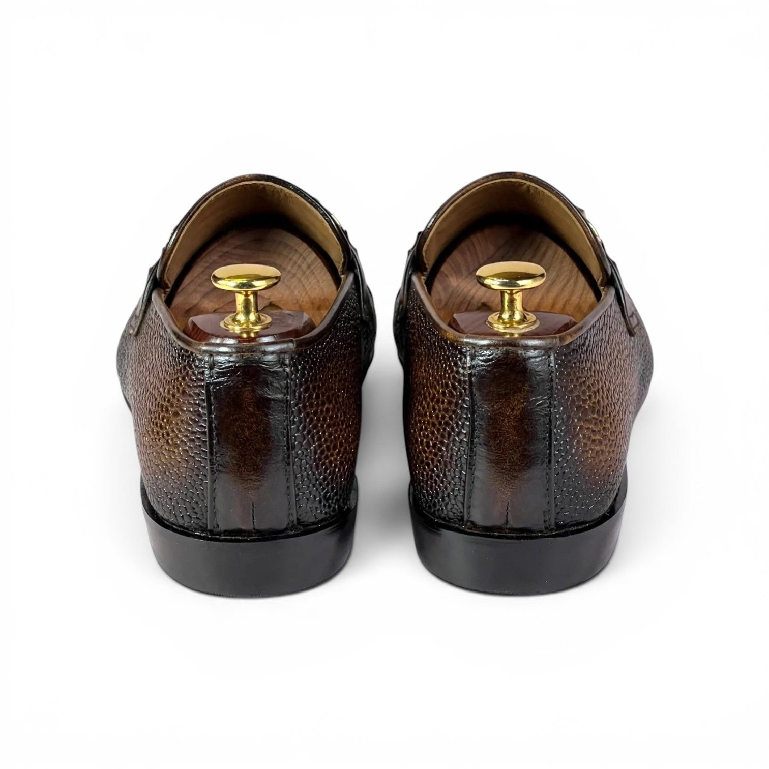 A pair of dark brown textured leather loafers shown from the back, fitted with wooden shoe trees and gold-colored metal pegs, on a plain white background.