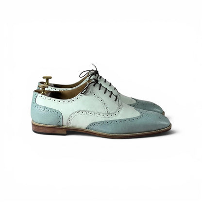 A pair of men's wingtip oxford shoes in a pale blue and white color-blocked design, shown on a white background with a wooden shoe tree inserted.