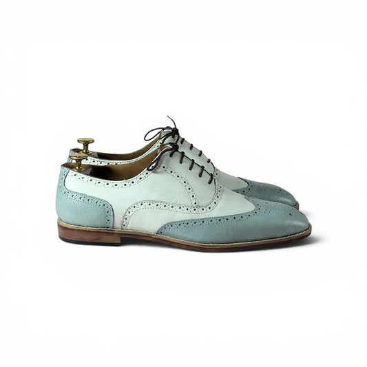 A pair of men's wingtip oxford shoes in a pale blue and white color-blocked design, shown on a white background with a wooden shoe tree inserted.
