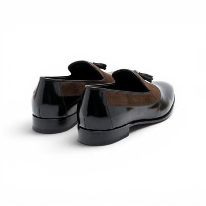 A pair of men's loafers featuring a glossy black patent leather toe cap and heel with a brown suede panel on the sides, finished with black tassels, shown from a rear three-quarter angle on a white background.