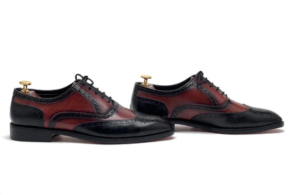 A pair of men's wingtip oxford shoes with a color-blocked design featuring black leather brogue toe caps and black laces, paired with dark red and black leather quarter panels. The shoes are displayed on wooden dress forms against a clean white background.