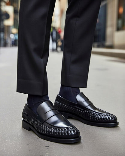 Black dress shoes with woven pattern worn with black pants on a blurred city street background