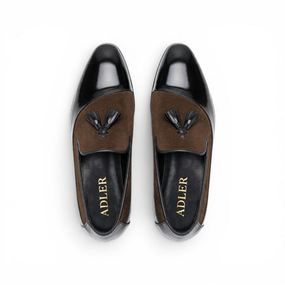 A pair of men's loafers shown from a top-down view on a white background. The shoes feature a glossy black leather toe cap and heel counter, a brown suede vamp with a black tassel, and a black insole with the brand name 'ADLER' in gold lettering.